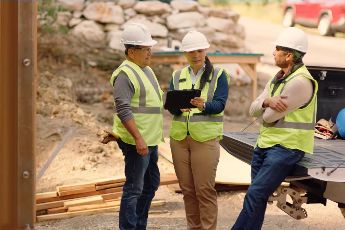 Three construction workers in safety vests and helmets discuss plans at a work site; one holds a tablet while another leans against a truck bed.