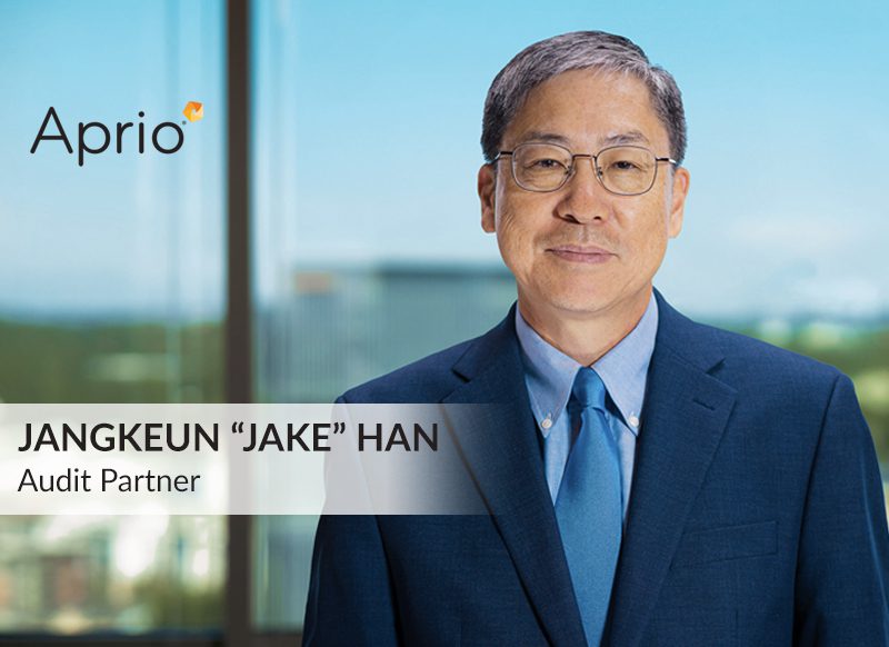 A man in a blue suit and glasses stands in an office with city views; text reads Jangkeun Jake Han, Audit Partner, Aprio.