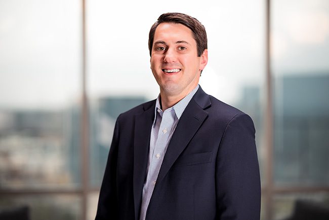 A man in a navy blazer and light blue shirt stands smiling in front of large windows with an out-of-focus cityscape in the background.