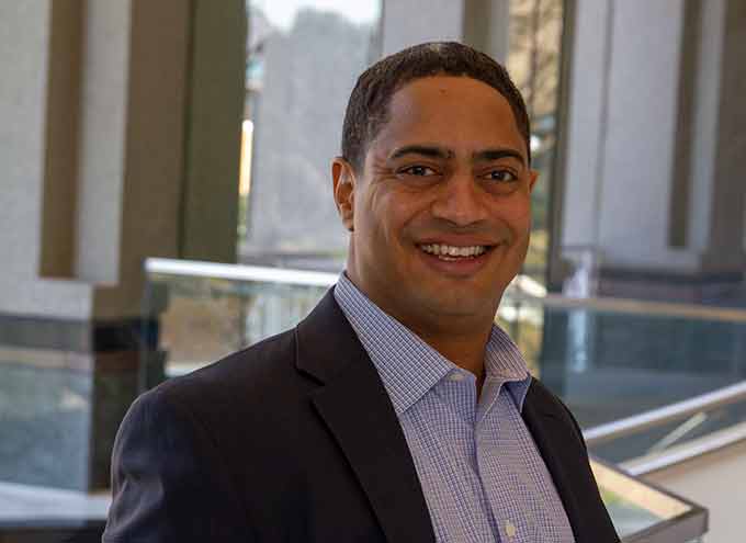 A man in a suit jacket and collared shirt stands indoors, smiling at the camera, with large windows and a glass railing in the background.