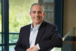 A man in a business suit smiles while leaning on a glass railing indoors, with large windows and greenery visible in the background.