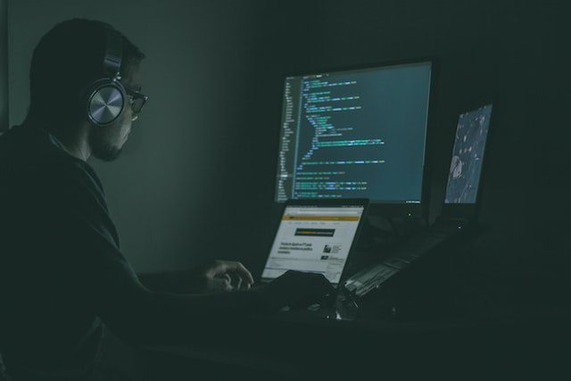 A person wearing headphones sits at a desk with three screens displaying code and a website, working in a dimly lit room.