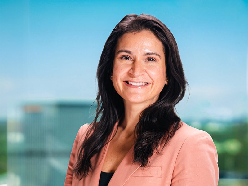 A woman with long dark hair, wearing a peach blazer over a black top, smiles at the camera with a blurred cityscape background.