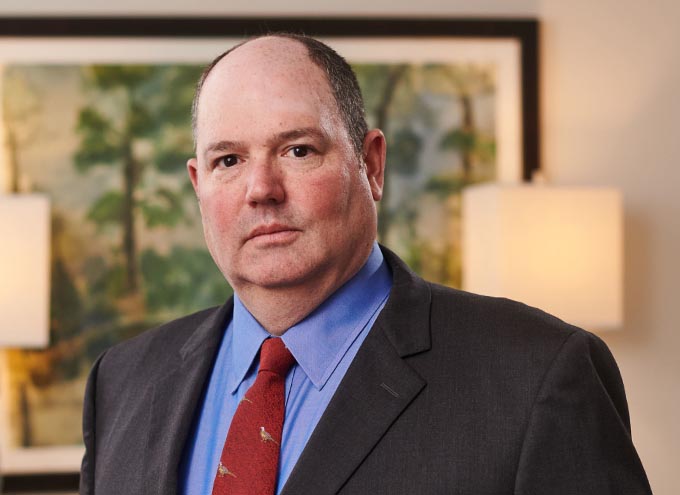 A man in a suit and red tie stands indoors in front of a blurred painting and two wall lights, facing the camera with a neutral expression.