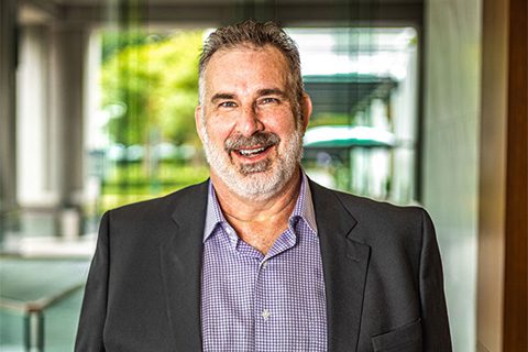 A middle-aged man with gray hair and a beard, wearing a dark blazer and purple checkered shirt, smiles while standing indoors with glass walls and greenery in the background.