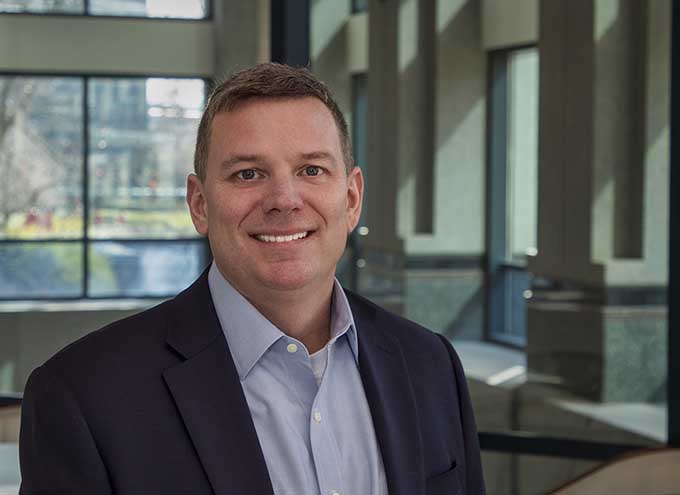Man in a blue shirt and dark blazer smiles while standing indoors near large windows with natural light.
