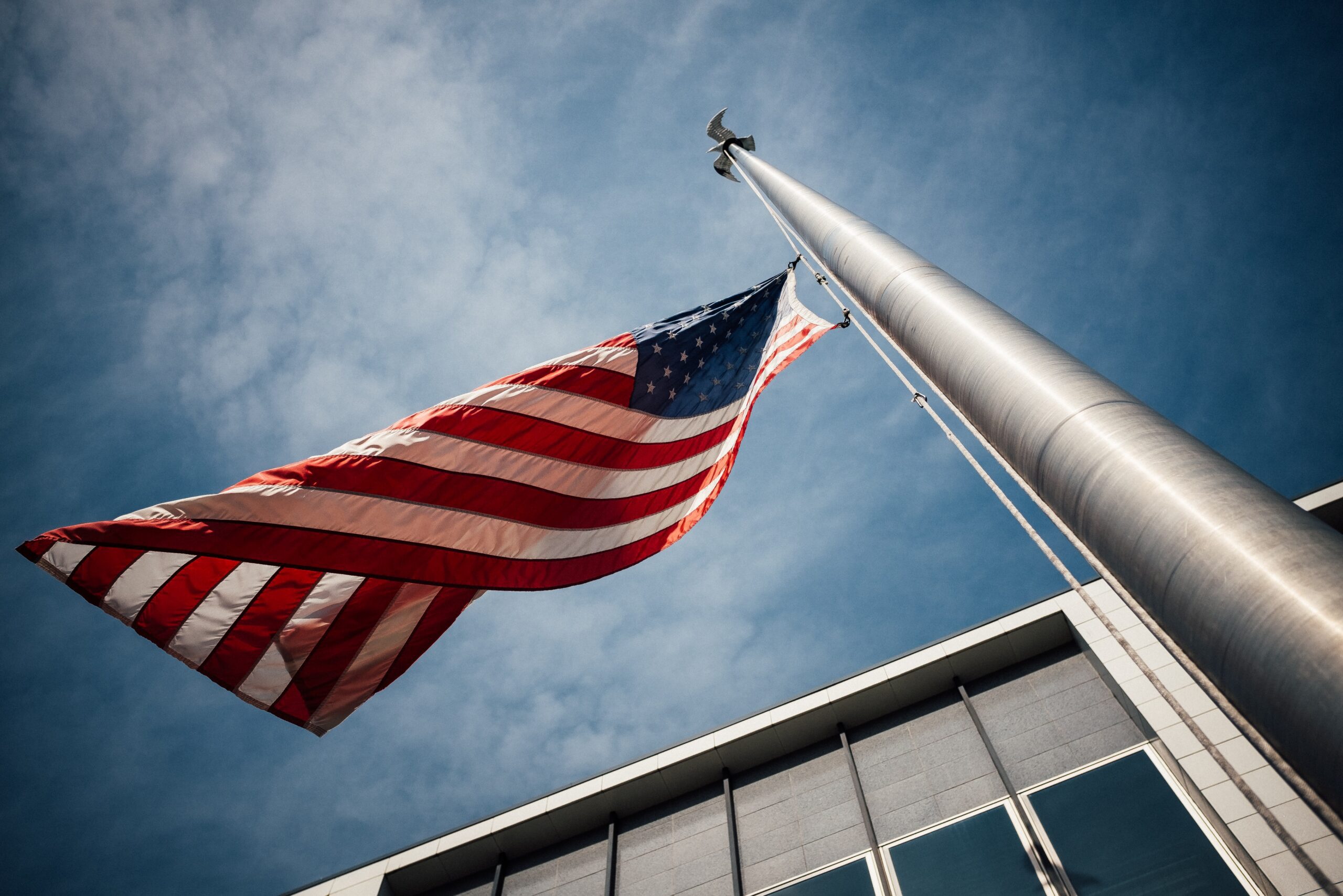 A United States flag waves on a flagpole outside a modern building, viewed from a low angle against a blue sky.