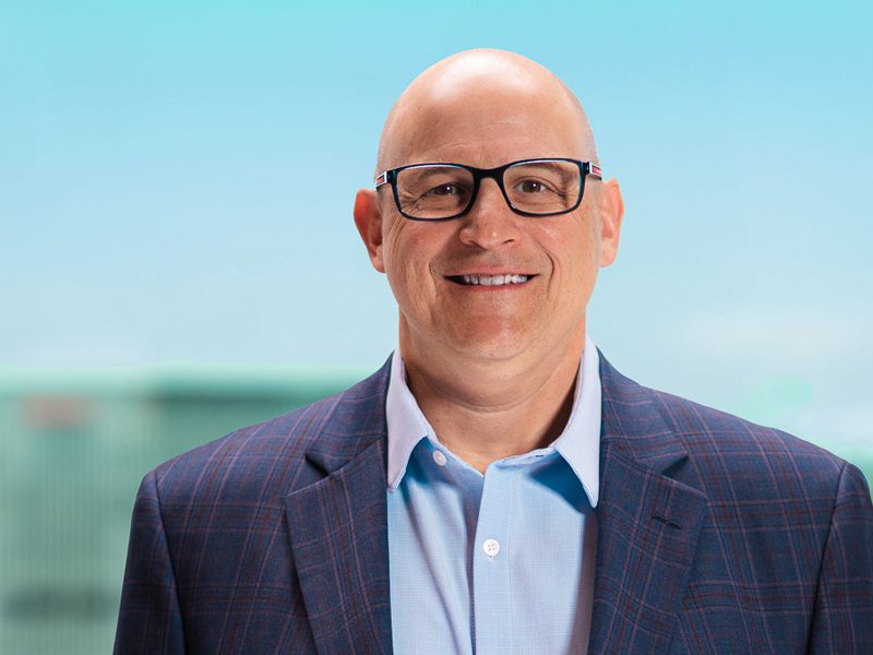 Bald man wearing glasses, a blue dress shirt, and a plaid blazer stands and smiles in front of a blurred outdoor background.