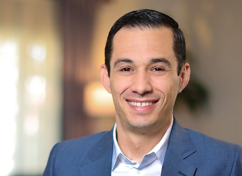 A man with short dark hair, wearing a blue suit jacket and light blue shirt, smiles at the camera in a softly lit indoor setting.