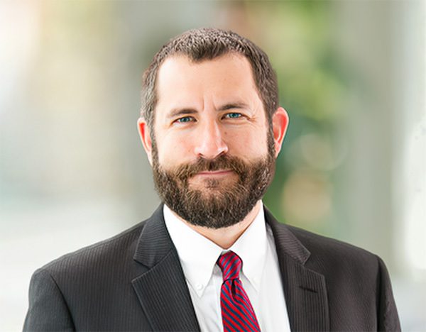 A man with a beard in a suit and red striped tie stands in front of a blurred background.