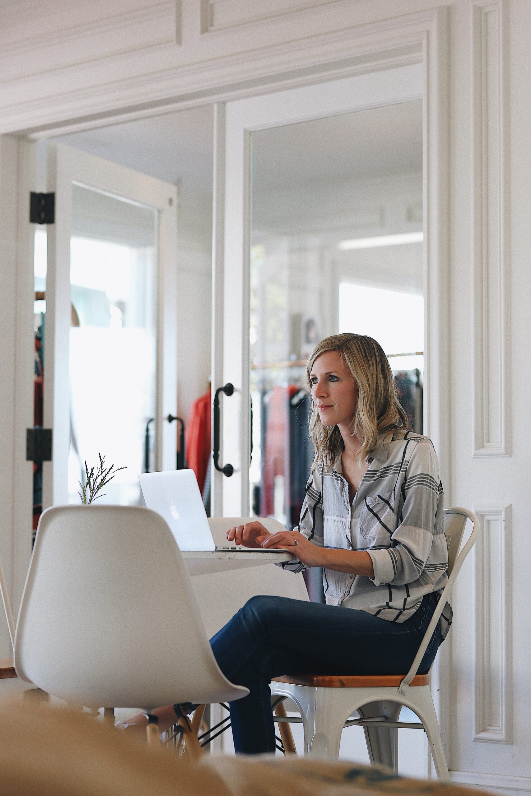 A woman sits at a desk using a laptop in a bright room with glass doors and a clothing rack visible in the background.