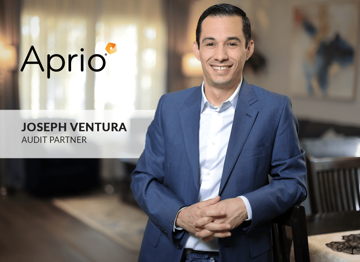 A man in a blue suit smiles at the camera with Aprio logo and text: Joseph Ventura, Audit Partner displayed beside him in a home office setting.
