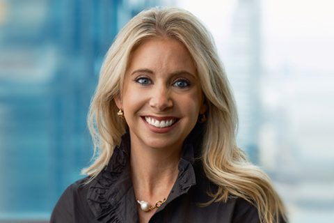 A woman with long blonde hair wearing a black top and a pearl necklace, smiling in front of a blurred office window background.