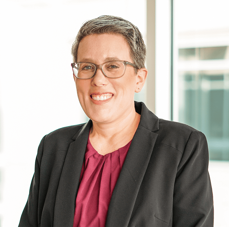 A person with short gray hair, glasses, and a dark blazer over a maroon blouse stands indoors, smiling in front of large windows.
