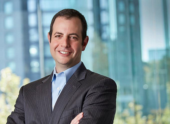 A man in a pinstripe suit stands with arms crossed, smiling, in front of a modern glass building.