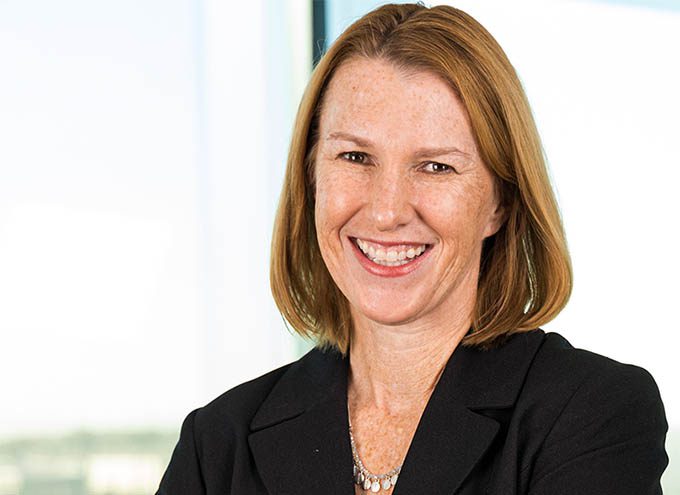 A woman with straight, shoulder-length auburn hair smiles, wearing a black blazer and a necklace, standing in front of a bright window.