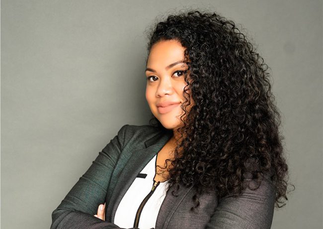 A woman with long curly hair, wearing a dark blazer and white top, stands with arms crossed against a plain grey background.