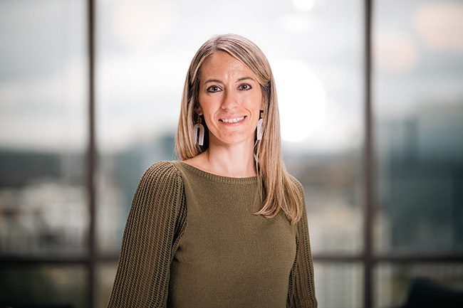 A woman with long blonde hair, wearing a green sweater and white earrings, stands indoors in front of large windows with a blurred cityscape background.