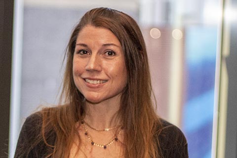 A woman with long brown hair, wearing a necklace and a dark top, stands indoors in front of a blurred background.