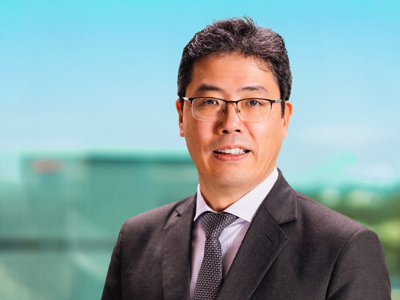 A man in a dark suit, light shirt, and patterned tie, wearing glasses, stands in front of a blurred outdoor background.
