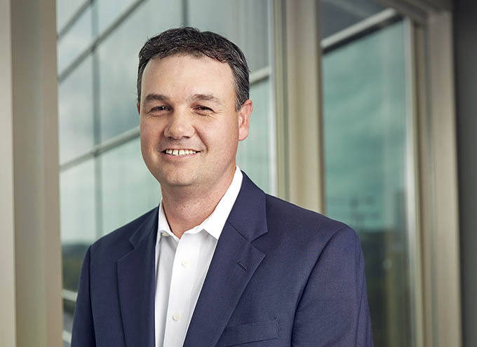 Man in a navy suit and white shirt standing indoors near large windows, smiling at the camera.