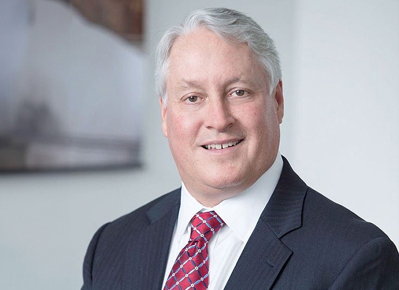 A man with gray hair wearing a dark suit, white shirt, and red patterned tie, standing in a bright indoor setting.