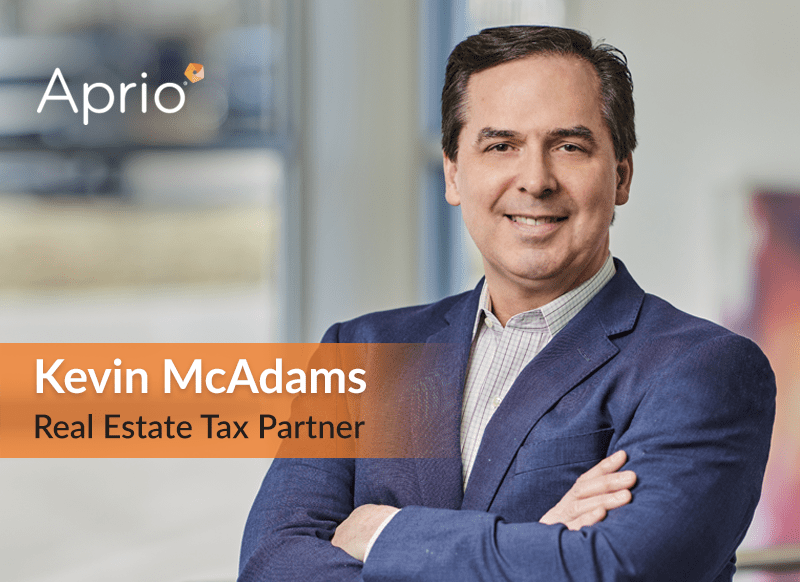 A man in a suit stands with arms crossed. Text reads: Aprio. Kevin McAdams, Real Estate Tax Partner. The background is an office setting.