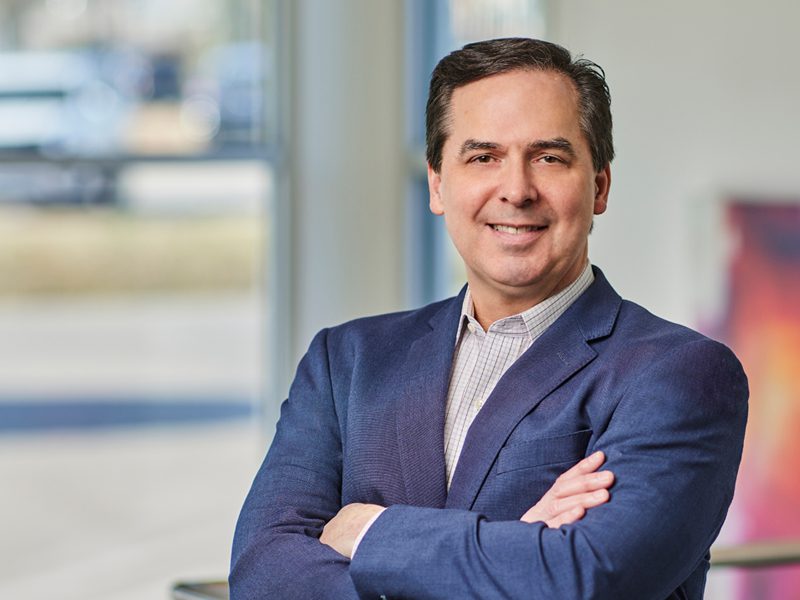 A man in a blue blazer with arms crossed stands indoors, smiling at the camera. A window and abstract artwork are visible in the background.
