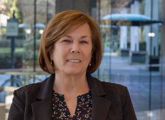 A woman with short brown hair wearing a black blazer and patterned blouse stands in front of large glass windows with an outdoor scene in the background.