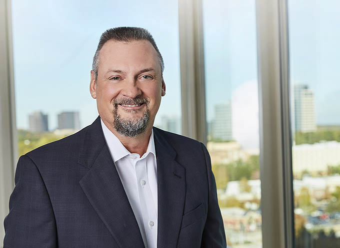 A man in a suit and white shirt smiles while standing in front of large windows with a cityscape visible in the background.