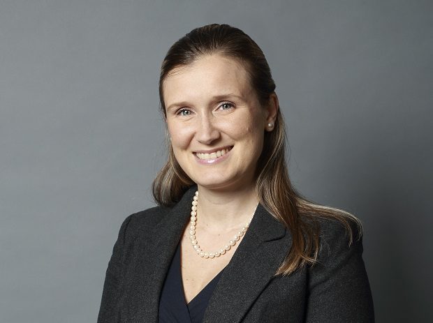 A woman with long brown hair, wearing a dark blazer, navy top, and pearl necklace, smiles at the camera against a plain gray background.