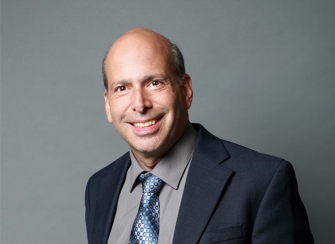 A middle-aged man with a bald head smiles at the camera. He is wearing a dark suit, gray shirt, and patterned blue tie against a plain gray background.