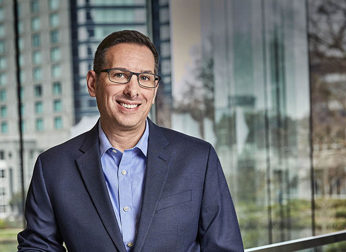 A man in a navy suit and glasses stands indoors in front of large windows with a cityscape in the background.