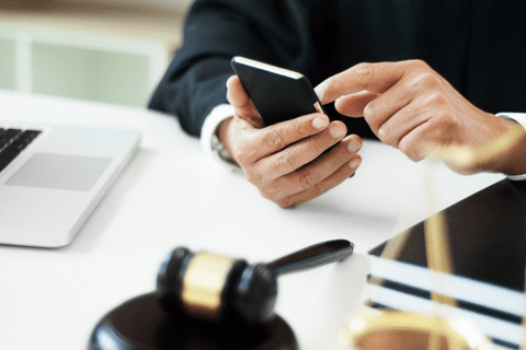 A person in professional attire uses a smartphone at a desk with a gavel, a notebook, and a laptop.
