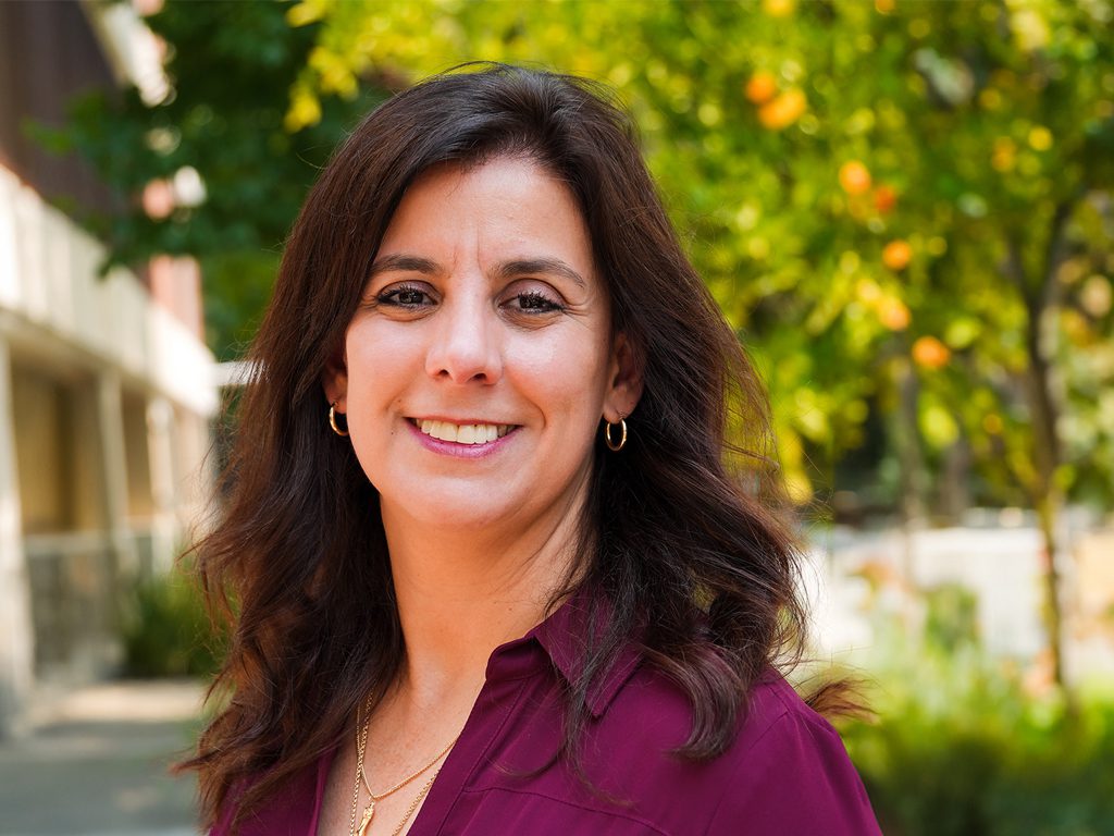 A woman with long dark hair wearing a burgundy blouse smiles outdoors with greenery and a building in the background.