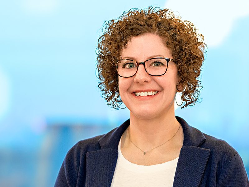 A woman with curly hair and glasses, wearing a blue blazer and white top, smiles while standing in front of a blurred blue background.