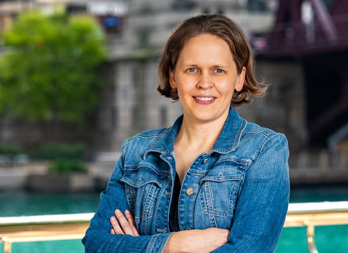 A woman with short brown hair wearing a blue denim jacket stands outdoors with her arms crossed, a river and cityscape in the background.