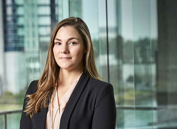 A woman with long brown hair wearing a black blazer and light blouse stands in front of large glass windows in a modern office setting.