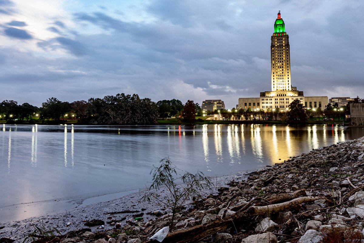 A tall, illuminated building stands beside a river at dusk with a rocky shoreline in the foreground and cloudy sky above.