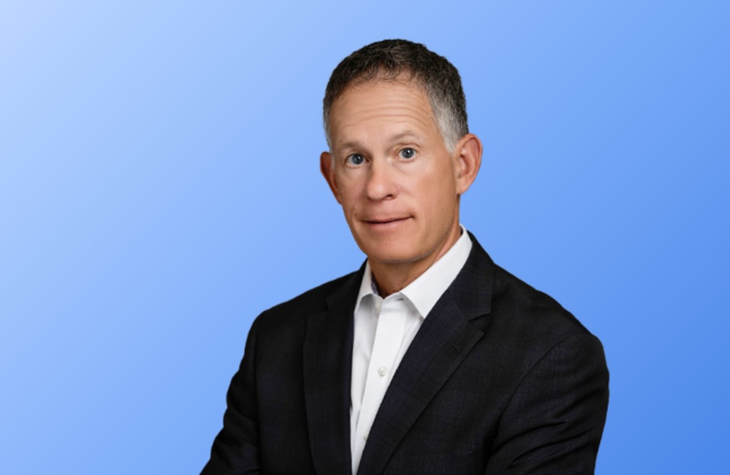 A man with short gray hair wearing a dark suit jacket and white dress shirt poses against a plain blue background.