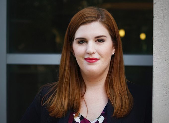 A woman with straight, shoulder-length auburn hair stands in front of a window, wearing a dark blazer and patterned top, looking at the camera with a neutral expression.
