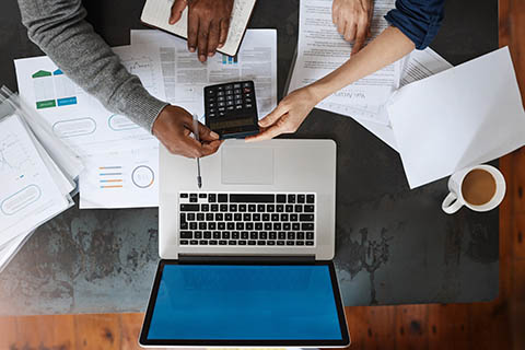 Two people work at a desk with a laptop, calculator, documents, a notebook, and a cup of coffee, viewed from above.