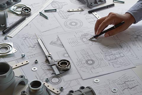 A person draws technical sketches on engineering blueprints surrounded by mechanical parts, calipers, and drafting tools on a desk.