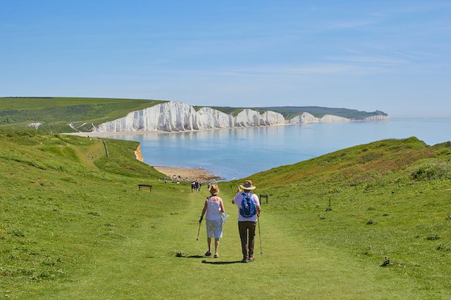 Two people walk along a grassy path toward white chalk cliffs by the sea on a clear, sunny day.