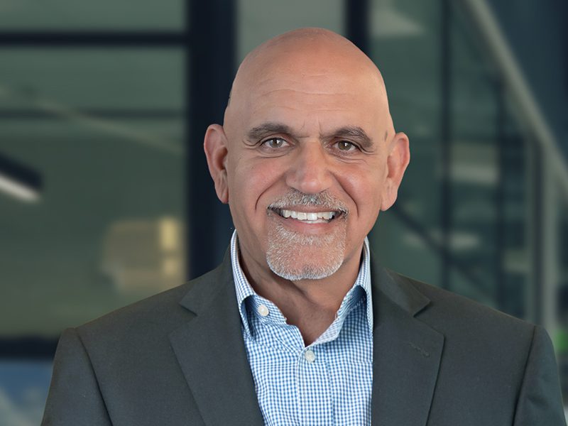 A bald man with a gray goatee, wearing a gray suit jacket and blue checked shirt, smiles in front of a blurred office background.