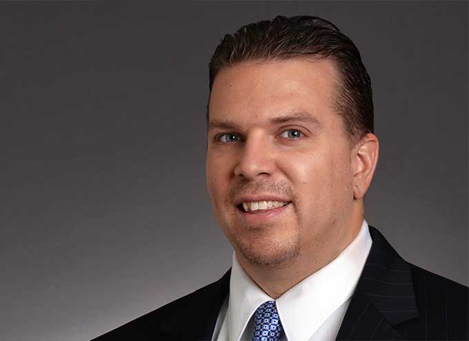 A man in a suit and tie poses for a professional headshot against a plain gray background.