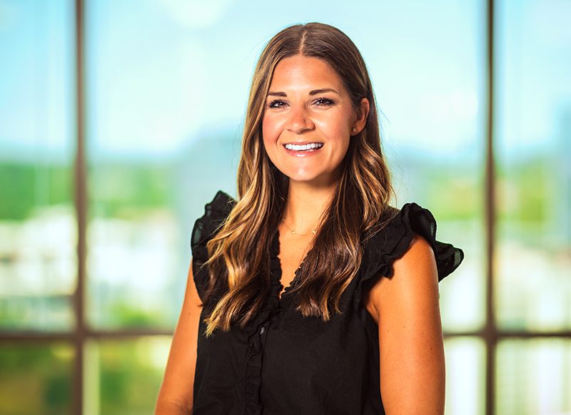 A woman with long brown hair, wearing a black sleeveless top, smiles while standing indoors in front of large windows with a blurred outdoor view.