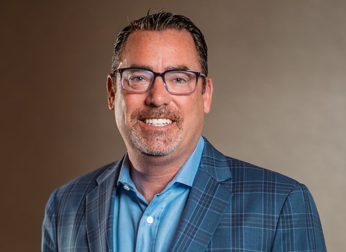 A man with short dark hair and glasses wearing a blue plaid suit jacket and light blue shirt smiles in front of a neutral background.