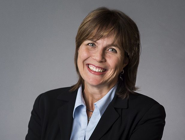 A woman with shoulder-length brown hair, wearing a black blazer and light blue shirt, smiles at the camera against a plain gray background.