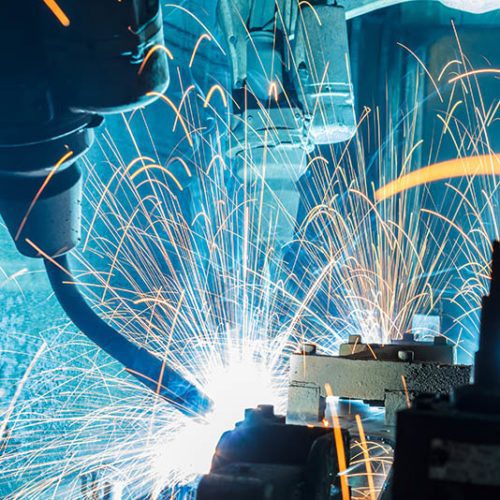 Robotic arms welding metal parts together, generating bright sparks and blue light in an industrial setting.